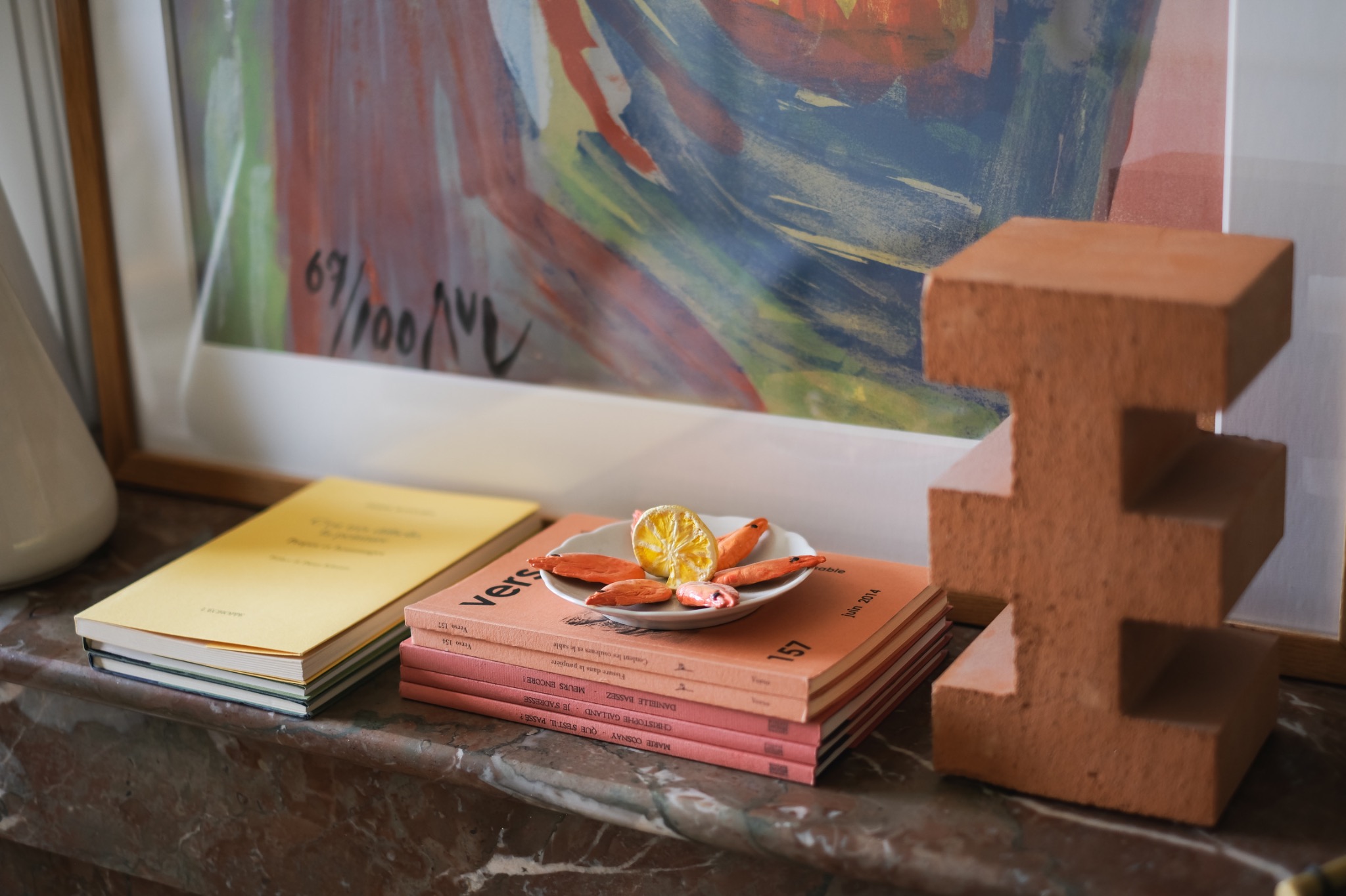 A curated material vignette with books, a small plate of sculptural objects, a framed artwork, and a terracotta architectural form.