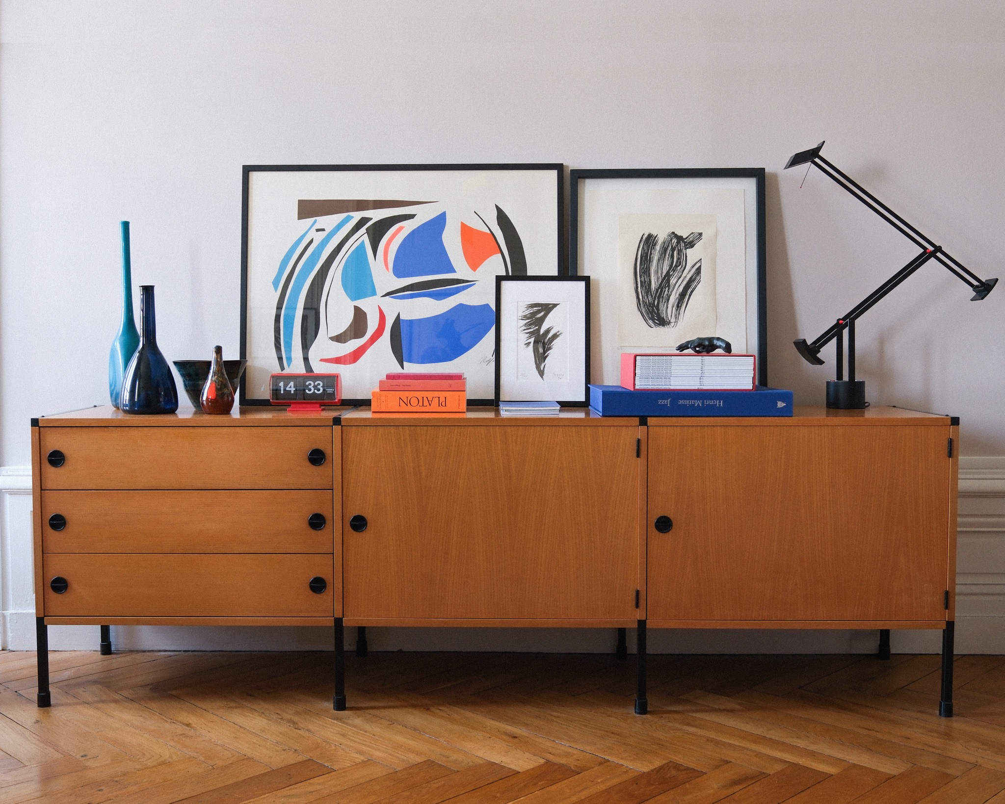 A refined interior vignette with a wooden sideboard, art, books, sculptural objects, and a black task lamp.